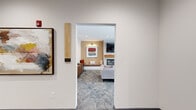 This single-floor communal space offers a well-appointed lounge environment designed for comfort and social engagement. The layout is organized into distinct yet interconnected zones, each serving a specific purpose while contributing to a cohesive, modern aesthetic.

The centerpiece of the space is a sitting room anchored by a sleek linear fireplace set against a white shiplap surround, topped by a wall-mounted television. Flanking the fireplace are built-in bench seats set beneath warm accent lighting, positioned against a textured chevron-patterned wallcovering in warm golden tones. Contemporary abstract artwork adorns the walls, adding visual interest and color. A sectional sofa and upholstered lounge chairs are arranged around a round wood-topped coffee table, creating an inviting focal point for gathering.

A secondary seating area near a row of windows offers lighter, more casual arrangements with individual chairs and small side tables, ideal for conversation or quiet relaxation. Roller shades allow for adjustable natural light throughout.

The space is finished with patterned low-pile carpeting throughout, recessed ceiling lighting, and clean white walls that enhance the brightness of the interior. An elevator and accessible entry corridor complete the layout, ensuring ease of movement throughout. The overall ambiance is warm, modern, and welcoming.
