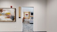 This single-floor communal space offers a well-appointed lounge environment designed for comfort and social engagement. The layout is organized into distinct yet interconnected zones, each serving a specific purpose while contributing to a cohesive, modern aesthetic.

The centerpiece of the space is a sitting room anchored by a sleek linear fireplace set against a white shiplap surround, topped by a wall-mounted television. Flanking the fireplace are built-in bench seats set beneath warm accent lighting, positioned against a textured chevron-patterned wallcovering in warm golden tones. Contemporary abstract artwork adorns the walls, adding visual interest and color. A sectional sofa and upholstered lounge chairs are arranged around a round wood-topped coffee table, creating an inviting focal point for gathering.

A secondary seating area near a row of windows offers lighter, more casual arrangements with individual chairs and small side tables, ideal for conversation or quiet relaxation. Roller shades allow for adjustable natural light throughout.

The space is finished with patterned low-pile carpeting throughout, recessed ceiling lighting, and clean white walls that enhance the brightness of the interior. An elevator and accessible entry corridor complete the layout, ensuring ease of movement throughout. The overall ambiance is warm, modern, and welcoming.