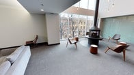 This elegantly designed single-floor space immediately impresses with its soaring, dramatic ceiling height and an expansive wall of floor-to-ceiling windows that floods the interior with natural light and offers open views of the surrounding urban streetscape. The generous proportions of the main lounge area create an airy, open ambiance that is both inviting and refined. A standout architectural feature is the striking freestanding cylindrical fireplace, finished in matte black, which serves as a bold focal point at the heart of the space. Set upon a circular raised platform, it adds a sculptural, mid-century modern sensibility to the room. Carefully selected furnishings — including iconic bentwood lounge chairs and warm wood accent pieces — complement the clean lines and neutral palette of the interior. The flooring features a plush, neutral-toned carpet that adds warmth and acoustic comfort underfoot. Crisp white walls contrast beautifully with the natural wood tones throughout, while recessed and pendant lighting provide a soft, layered glow. The overall ambiance is calm, sophisticated, and thoughtfully curated, making this a welcoming and visually engaging space suited for relaxed gathering or quiet contemplation.