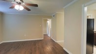 This single-story space presents a well-maintained interior with a cohesive, understated aesthetic. The main living area is finished with rich, dark hardwood-style flooring that runs throughout, providing a warm and grounded feel underfoot. Crown molding lines the upper perimeter of the walls, adding a subtle architectural detail that elevates the overall finish of the space. A ceiling fan is centrally positioned, offering both practical air circulation and a functional overhead fixture.

The walls are painted in a soft, neutral warm beige tone, creating a welcoming atmosphere that pairs well with the darker flooring. Crisp white baseboards provide clean contrast along the floor line. Visible HVAC venting integrated into the walls suggests a central air system throughout.

A doorway leads into a connecting hallway, which is illuminated by a wall-mounted light fixture, hinting at additional rooms beyond. The passageway is bright and open, suggesting a straightforward and accessible floor plan. The overall ambiance of the space is calm and neutral, offering a versatile foundation well-suited for a variety of interior styles and furnishing arrangements.