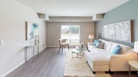 This single-floor residence offers a thoughtfully designed open-concept layout connecting the living and kitchen areas. The living room is spacious and bright, featuring wood-look flooring, large windows with outdoor views of greenery, and a distinctive accent wall in a deep gray tone that adds visual depth. Structural beam details along the ceiling contribute architectural character throughout.

The kitchen is well-equipped with rich dark-wood cabinetry, a classic white subway tile backsplash, stainless steel appliances including a refrigerator, range, microwave, and dishwasher, as well as a central island with bar seating and light quartz countertops. Track lighting illuminates the space effectively.

The bedrooms feature soft, neutral-toned carpeting and windows that allow natural light to filter in, with closet access visible in each room. The primary bathroom includes a double-sink vanity with dark cabinetry, a white countertop, a large mirror, and stone-look tile flooring. A secondary bathroom offers a single-sink vanity with similarly refined finishes. The overall palette throughout the unit is neutral and cohesive, creating a calm, modern ambiance.