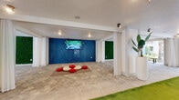 Located at 13645 E Colonial Dr in Orlando, FL, this single-floor amenity space is designed with a contemporary aesthetic that blends vibrant interior design with inviting outdoor elements. The interior is anchored by a striking cobalt-blue herringbone-patterned tile accent wall, which serves as a dramatic backdrop for a mounted flat-screen display and casual floor-level seating with colorful cushions. Adjacent lounge areas feature bold vertical dark wood slat accent walls, contemporary sculptural blue chairs, and patterned area rugs, creating intimate yet stylish gathering spots. Lush green moss walls add a natural, organic texture throughout the space, while draping white curtains provide an airy, flexible division between zones. Large tile flooring runs consistently throughout, giving the space a clean, cohesive look. The interior opens directly to a serene landscaped courtyard framed by tall palm trees, native plantings, and stone gravel beds, offering residents a tranquil outdoor retreat. Paved walkways and bench seating make the courtyard a functional extension of the interior. Safety features including fire suppression equipment are thoughtfully integrated. Overall, the space delivers a well-curated balance of modern design, comfort, and community functionality.