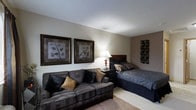 Located at 300 Springboro Lane in Columbus, Ohio, this single-floor space offers a thoughtfully arranged layout that combines comfort and functionality. The main living and sleeping area is designed with a cohesive aesthetic, featuring neutral warm tones on the walls that create a calm and inviting ambiance. A plush, dark upholstered sofa anchors the seating area, accented with decorative throw pillows in complementary earth tones, providing a relaxed space for lounging. Positioned adjacent to the sofa, a dark-finished bed frame with coordinating bedding integrates seamlessly into the room, making efficient use of the available space. A bedside nightstand with a classic table lamp adds a soft, warm glow to the room, enhancing its cozy atmosphere. The walls are adorned with framed botanical artwork, adding a refined, decorative touch, while a textured wall sculpture introduces an element of visual interest. The flooring transitions from hard surface to plush carpet, further defining the living and sleeping zones. The overall design presents a clean, well-appointed interior with consistent dark wood furnishings and carefully chosen accents that give the space a polished, comfortable feel.