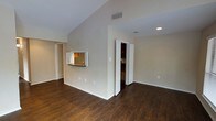 This single-floor space is defined by its vaulted ceilings, which lend a sense of openness and volume to the main living area. The floors throughout are finished in rich, dark hardwood-style planks, providing a warm and cohesive aesthetic that contrasts cleanly with the neutral beige walls and bright white trim.

A notable architectural feature is the pass-through window connecting the living area to the kitchen, framed with white shutters and accented by a dark countertop ledge. This design element maintains visual connectivity between spaces while preserving a degree of separation. The kitchen, visible through the pass-through, features warm-toned cabinetry and appears well-lit.

Recessed lighting in the ceiling supplements the natural brightness of the space, ensuring even illumination throughout. A bi-fold door is visible along one wall, likely accessing a closet or utility area, adding practical storage to the layout.

The clean lines, high ceilings, and open floor plan create a versatile living environment that feels both spacious and functional, suitable for a variety of furniture arrangements and uses.