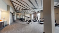 This single-level fitness center is designed to provide a comprehensive workout environment within a spacious, open layout. The room's most striking architectural feature is its coffered ceiling, accented with dark beam detailing that adds visual depth and a polished aesthetic to the space. Neutral-toned walls and gray carpeted flooring contribute to a clean, modern atmosphere throughout.

The cardio zone lines the perimeter of the room, where large floor-to-ceiling windows flood the space with natural light and offer views of the surrounding outdoor area, including a pool and neighboring buildings. Multiple treadmills, elliptical machines, and stationary bikes are positioned to take full advantage of these views, making workouts more engaging.

The strength training area features cable machines, a power rack, adjustable benches, and a dumbbell station with wall-mounted mirrors that visually expand the space and allow users to monitor their form. Stability and exercise balls are stored along one wall, supporting flexibility and functional fitness routines.

Near the entrance, a well-organized service area provides sanitation supplies, storage cubbies, and cabinetry, ensuring the space remains tidy and functional. Ceiling fans throughout aid in air circulation, enhancing comfort during intense workouts. Overall, the fitness center balances practicality with thoughtful design.