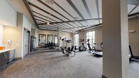 This single-level fitness center is designed to provide a comprehensive workout environment within a spacious, open layout. The room's most striking architectural feature is its coffered ceiling, accented with dark beam detailing that adds visual depth and a polished aesthetic to the space. Neutral-toned walls and gray carpeted flooring contribute to a clean, modern atmosphere throughout.

The cardio zone lines the perimeter of the room, where large floor-to-ceiling windows flood the space with natural light and offer views of the surrounding outdoor area, including a pool and neighboring buildings. Multiple treadmills, elliptical machines, and stationary bikes are positioned to take full advantage of these views, making workouts more engaging.

The strength training area features cable machines, a power rack, adjustable benches, and a dumbbell station with wall-mounted mirrors that visually expand the space and allow users to monitor their form. Stability and exercise balls are stored along one wall, supporting flexibility and functional fitness routines.

Near the entrance, a well-organized service area provides sanitation supplies, storage cubbies, and cabinetry, ensuring the space remains tidy and functional. Ceiling fans throughout aid in air circulation, enhancing comfort during intense workouts. Overall, the fitness center balances practicality with thoughtful design.