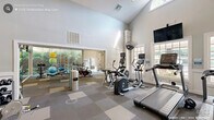 Located in Raleigh, NC, this well-appointed community fitness center offers a comprehensive workout and relaxation destination across a single floor. The main gym area is the centerpiece of the facility, showcasing soaring vaulted ceilings with large multi-pane windows that flood the space with natural light and offer views of the surrounding greenery. The gym is outfitted with a wide array of equipment, including treadmills, ellipticals, stationary and cycling bikes, a multi-station strength machine, a TRX suspension system, free weights, and a workout bench, all arranged across a durable checkered tile floor. Interior glass partitions divide workout zones while maintaining an open, airy feel. A connected lounge area provides a welcoming retreat, featuring warm hardwood-style flooring, a large wall-mounted TV, comfortable seating including a caramel leather sofa and accent chairs, a work table with natural light from oversized windows, and tasteful wall art. A dedicated coffee station with a tile-accented accent wall and a selection of single-serve options adds a convenient amenity for residents. The overall ambiance is modern and clean, with a neutral color palette, recessed lighting, and a cohesive design that balances functionality with comfort.