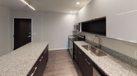 This thoughtfully designed single-floor residence showcases a clean, contemporary aesthetic with consistent finishes throughout. The open-concept kitchen is a standout feature, equipped with stainless steel appliances including a range, microwave, refrigerator, and dishwasher. Dark lower cabinetry is paired with crisp white upper cabinets, while speckled granite countertops extend across both the perimeter and a central island, providing generous prep and storage space. Recessed lighting illuminates the kitchen, which flows seamlessly into the open living area finished with rich, dark hardwood-style flooring. The bedroom offers a comfortable, well-proportioned layout with a large window that brings in natural light and urban views. A dedicated hallway connects the bedroom to additional rooms, including a closet and a full bathroom. The bathroom features a granite-topped dark vanity with an undermount sink, a wall-mounted mirror, and a tub-shower combination with clean white tile surround. A dedicated laundry room houses a full-size washer and dryer alongside overhead wire shelving for added storage. Neutral wall tones, white trim, and consistent flooring create a cohesive and modern atmosphere throughout the entire space.