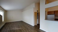 This single-floor space offers a clean and functional layout characterized by a bright, neutral color palette and durable dark hardwood-style flooring that runs throughout the main living area. The room benefits from vaulted ceilings that lend an open, airy quality, enhancing the overall sense of space and volume. White walls provide a versatile backdrop, allowing the architectural lines of the angled ceiling to stand out as a defining design feature. A wooden bi-fold closet door is visible at the far end of the room, offering built-in storage conveniently integrated into the floor plan. The HVAC vent positioned near the ceiling ensures comfortable climate control year-round. The layout is straightforward and efficient, providing a flexible living area that can be adapted to a variety of furniture arrangements. The combination of clean lines, neutral finishes, and practical storage solutions gives the space a move-in-ready appeal with a simple, understated aesthetic throughout.