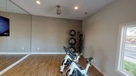 This single-floor space is purpose-built as a private fitness room, designed to support a well-rounded workout routine. The room features warm-toned, wood-look flooring that provides both durability and a clean aesthetic underfoot. Neutral taupe walls create a focused, distraction-free environment conducive to exercise.

Overhead, recessed lighting provides consistent, even illumination throughout the space, complemented by a ceiling-mounted light fixture that adds an additional layer of brightness. The ceiling height is comfortable, allowing ample room for standing and overhead movements.

The fitness area is outfitted with multiple spin bikes positioned side by side, making it suitable for solo cycling sessions or group workouts. Along one wall, a vertical storage rack holds several stability balls of varying sizes, keeping the floor clear and the equipment organized. An adjacent rack stores resistance bands, medicine balls, and other accessories, ensuring a variety of training options are readily accessible.

Clean white baseboards frame the perimeter of the room, adding a polished finish to the overall design. The layout is open and functional, offering enough floor space for stretching, floor exercises, or additional equipment. Overall, the space is thoughtfully arranged to deliver a comprehensive and efficient home fitness experience.