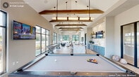 Located in Cary, NC, this sophisticated club lounge is a single-floor amenity space designed for both relaxation and entertainment. The interior is anchored by dramatic vaulted ceilings adorned with exposed wood beams and elegant pendant lighting, creating a sense of grandeur throughout the open layout. Light hardwood flooring and large black-framed windows flood the space with abundant natural light while offering views of the surrounding community.

The game area serves as a central focal point, featuring a full-size billiards table and a shuffleboard table, both complemented by bold geometric area rugs. The adjoining living area includes plush navy blue sofas, a marble coffee table, and curated wall art, all bathed in natural light from floor-to-ceiling windows.

The kitchen and bar area is a standout, showcasing powder blue cabinetry, a dramatic marble backsplash, open wood shelving with gold accents, globe pendant lights, and a sweeping curved quartz island with upholstered barstool seating. A wall-mounted TV completes the entertainment setup.

A covered outdoor deck with ceiling fan and seating extends the living space outside, offering a relaxed retreat. The overall design blends modern elegance with casual comfort, making it an inviting communal destination.