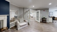 Located at 13430 Coppermine Rd in Herndon, Virginia, this single-floor space comprises a living room and a dedicated office, offering a thoughtfully designed environment that balances comfort with functionality. The living room is a standout feature, lined with expansive white built-in bookshelves that stretch across multiple walls, housing an extensive collection of books. A round dining-style table with upholstered white chairs sits at the heart of the library area, creating an inviting space for reading or conversation. A cozy seating nook anchors one corner, complete with a classic white fireplace framed by a bold navy blue accent wall, warm framed artwork, and an upholstered armchair — ideal for quiet relaxation. Elegant crown molding, recessed lighting, and neutral gray walls throughout lend the space a polished, refined aesthetic. Wide-plank wood-look flooring flows seamlessly across the entire floor, adding warmth and continuity. White French doors connect the rooms, enhancing the open and airy feel. The office area is equipped with a clean-lined white workstation, ergonomic seating, and additional accent chairs, making it a practical and comfortable workspace. Together, the rooms create a cohesive, welcoming environment with a sophisticated residential character.