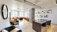 Located at 4899 Montrose Boulevard in Houston's vibrant Montrose neighborhood, this single-floor communal lounge is a thoughtfully designed social space that blends contemporary aesthetics with functional comfort. The open-plan layout is unified by warm travertine tile flooring and crisp white walls, creating a bright and welcoming atmosphere throughout.

The space is anchored by a generously sized living area featuring a plush sectional sofa, tan leather club chairs, and multiple small-group seating arrangements centered around bistro-style tables — ideal for both casual lounging and informal gatherings. Tall, floor-to-ceiling windows flood the interior with natural light and offer expansive views of the surrounding Houston skyline.

Architectural character is provided by exposed concrete columns and a partially exposed ceiling with industrial-style ductwork, balanced by refined track lighting that highlights the curated art collection adorning the walls. A gallery-style arrangement of black-and-white framed prints adds a sophisticated focal point near the dining counter area, which features a sleek dark wood high-top table and bar seating alongside a marble-topped reception counter.

Additional seating nooks with woven accent chairs and round mirrors contribute to the layered, gallery-like ambiance. A large, colorful abstract painting further enlivens the lounge zone, making this space as visually engaging as it is inviting.