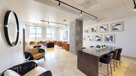 Located at 4899 Montrose Boulevard in Houston's vibrant Montrose neighborhood, this single-floor communal lounge is a thoughtfully designed social space that blends contemporary aesthetics with functional comfort. The open-plan layout is unified by warm travertine tile flooring and crisp white walls, creating a bright and welcoming atmosphere throughout.

The space is anchored by a generously sized living area featuring a plush sectional sofa, tan leather club chairs, and multiple small-group seating arrangements centered around bistro-style tables — ideal for both casual lounging and informal gatherings. Tall, floor-to-ceiling windows flood the interior with natural light and offer expansive views of the surrounding Houston skyline.

Architectural character is provided by exposed concrete columns and a partially exposed ceiling with industrial-style ductwork, balanced by refined track lighting that highlights the curated art collection adorning the walls. A gallery-style arrangement of black-and-white framed prints adds a sophisticated focal point near the dining counter area, which features a sleek dark wood high-top table and bar seating alongside a marble-topped reception counter.

Additional seating nooks with woven accent chairs and round mirrors contribute to the layered, gallery-like ambiance. A large, colorful abstract painting further enlivens the lounge zone, making this space as visually engaging as it is inviting.