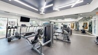 Located at 900 Downtowner Boulevard in Mobile, Alabama, this well-appointed fitness center offers a comprehensive workout environment within a single, efficiently organized space. The room features a striking tray ceiling finished in a deep charcoal tone, complemented by bright linear LED lighting and recessed fixtures that ensure thorough illumination throughout. Large windows line the walls, inviting ample natural light and offering views of the surrounding greenery, creating an open and energizing atmosphere. Floor-to-ceiling mirrors span a full wall, visually expanding the space and allowing users to monitor their form during exercise. The cardio section is equipped with a lineup of TRUE-brand treadmills, elliptical trainers, and a stationary bike, providing a range of aerobic workout options. The strength training area includes a cable machine, a leg press/extension machine, and additional selectorized equipment from TRUE Fitness, alongside a well-stocked dumbbell rack with a flat weight bench for free weight exercises. Durable rubber flooring covers the entire area, providing safety and comfort underfoot. A wall-mounted flat-screen television adds entertainment. The overall design is clean, modern, and functional, making this fitness center a practical and well-equipped facility for a full-body workout routine.
