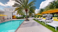This single-level outdoor space offers a resort-style atmosphere centered around a shimmering swimming pool with crisp turquoise water. The pool area is framed by mature, towering palm trees that provide natural shade and a distinctly tropical ambiance. A paved deck surrounds the pool, finished in interlocking pavers that provide a clean and modern aesthetic underfoot. Arranged along the deck are sleek, contemporary lounge chairs dressed in bold yellow cushions and pillows, inviting relaxation in the sun. A lush green lawn extends alongside the pool, offering additional open space for leisure. Vibrant yellow market umbrellas provide shade over additional seating areas, complementing the bright and energetic color palette of the space. A pergola structure is visible in the background, creating a shaded gathering zone with outdoor furniture. Dense tropical greenery and manicured hedges line the perimeter, offering privacy and a sense of seclusion. The surrounding architecture features clean white modern facades, enhancing the bright, sun-drenched character of the environment. A colorful accent wall with bold artwork adds a lively visual element to the space. String lights are strung across the pergola area, suggesting a warm and inviting atmosphere for evening use as well. Overall, this outdoor space combines modern design with lush tropical surroundings.