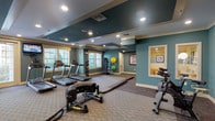 This fitness center offers a comprehensive workout environment designed to accommodate a wide range of exercise routines. The space is finished with teal-painted walls, cream-colored trim, and a distinctive tray ceiling with a dark beadboard inset, lending the room a polished, upscale aesthetic. Recessed lighting throughout ensures bright, even illumination across the entire floor.

The cardio section is well-appointed with multiple Matrix treadmills positioned in front of large windows fitted with horizontal blinds, allowing natural light to filter in while providing views of outdoor greenery. A wall-mounted flat-screen television serves the cardio area, keeping exercisers entertained during workouts. Alongside the treadmills, a row of elliptical machines lines a mirrored wall, which visually expands the space and allows users to monitor their form.

A dedicated functional training zone features heavy-duty equipment including battle ropes, a large tire for flipping drills, and a sled, offering options for high-intensity interval training. Stationary cycling bikes are positioned in their own area, accompanied by a storage unit. Exercise balls in multiple sizes are neatly organized on a rack in the corner. The flooring consists of durable, textured carpet throughout, providing comfort and traction. Additional rooms visible through interior windows suggest supplementary spaces such as a stretching or studio area.
