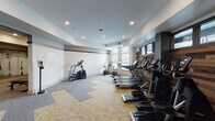 This fitness center is thoughtfully designed to provide a comprehensive workout environment within a single, well-organized floor. The main cardio area is expansive and airy, featuring a row of treadmills, elliptical trainers, and stationary bikes aligned along a striking reclaimed wood accent wall. A band of clerestory windows positioned above the wood paneling allows natural light to filter in, complementing the recessed ceiling lighting that keeps the space bright and inviting. The patterned carpet tiles in neutral grey and yellow tones add a modern, energetic aesthetic underfoot while offering comfort during exercise. A dedicated strength and functional training room adjoins the main cardio area, equipped with adjustable benches and TRX suspension training straps mounted on a wall-length rack. Full-length mirrors with yellow-trimmed frames line one wall of this room, enhancing the sense of space and allowing users to monitor their form. The entrance corridor features warm hardwood flooring and clean white walls adorned with framed abstract artwork in black and gold tones, setting an upscale tone upon arrival. Dark wood door frames and trim throughout tie the design together cohesively, creating a polished and modern fitness facility.