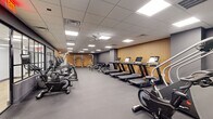 This modern fitness center offers a comprehensive workout environment designed to accommodate a wide range of exercise needs. The main floor is organized into distinct functional zones, providing ample space for both cardio and strength training activities. A row of treadmills lines one wall, backed by a striking vertical wood slat accent panel that adds warmth against the predominantly grey and charcoal color palette. Ceiling fans and recessed panel lighting ensure consistent air circulation and even illumination throughout. The cardio zone also features elliptical trainers, rowing machines, and stationary bikes arranged to maximize floor space and ease of movement. A dedicated strength training area hosts a variety of selectorized cable machines and a squat rack, offering a full range of resistance training options. A standout feature is a separate enclosed studio room, defined by elegant black-framed glass partition walls that maintain an open visual connection to the main floor. This studio is equipped with a mounted display screen and spin bikes, suitable for group classes or personal training sessions. Durable rubber flooring covers the entire facility, providing both safety and functionality. The overall design is clean and contemporary, with thoughtful material choices — including the warm wood paneling and soft mint-toned studio walls — creating an inviting and motivating atmosphere.