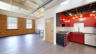 Located at 230 Scott Street in Buffalo, NY, this 2-bedroom, 1-bathroom loft blends industrial character with modern finishes across a single thoughtfully laid-out floor. The open living area makes a strong first impression with soaring ceilings, exposed structural beams, and large-diameter ductwork that celebrate the building's industrial heritage. Original brick accent walls line the living space, complemented by wide-plank hardwood-style flooring and generous windows that invite natural light and street views. A sleek kitchen features bold red high-gloss cabinetry paired with dark countertops and a stainless steel gas range, creating a striking visual contrast. A modern black steel railing delineates the split-level transition within the space, adding an architectural focal point. The expansive primary bedroom is carpeted and features tall ceilings with exposed infrastructure, offering a sense of openness, along with access to closet space. The second bedroom is similarly sized and carpeted. The bathroom is finished with tile flooring, a granite-topped vanity with under-cabinet storage, and a large mirror. Throughout, the space balances raw industrial elements with clean, contemporary touches.