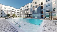 Located at 10850 Hammerly in Houston, Texas, this outdoor amenity space is thoughtfully designed around a central resort-style swimming pool that serves as the focal point of the courtyard. The multi-story building wraps around the space in a sweeping U-shaped configuration, finished in a contemporary palette of gray and white horizontal lap siding with vertical board accents, creating a cohesive and modern aesthetic.

The expansive pool deck is surfaced with light-toned concrete and decorative paver walkways featuring a geometric pattern, providing both visual interest and durable all-weather functionality. A generous row of lounge chairs lines the poolside, offering ample space for sunbathing beneath the open Texas sky. Water features at the pool's edge add a dynamic element to the serene setting.

Adjacent to the pool, a covered pergola structure provides shaded outdoor dining with a built-in grill station, making it an ideal space for social gatherings. Nearby seating areas feature stylish wicker chairs and contemporary furnishings arranged around curated garden beds planted with ornamental grasses, flowering shrubs, and young shade trees that add greenery and color throughout the courtyard.

Gated entry points with decorative metal fencing and paver pathways lead into the space, enhancing both security and curb appeal. The overall ambiance is bright, open, and resort-inspired, offering a welcoming outdoor retreat.