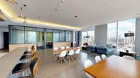Located at 615 W Division St in Chicago's vibrant Near North Side, this sophisticated amenity space combines contemporary design with a warm, functional atmosphere. The open-plan layout is anchored by expansive floor-to-ceiling windows that flood the interior with natural light and frame breathtaking panoramic views of the Chicago skyline and surrounding neighborhoods. A generously sized lounge area serves as the social heart of the space, furnished with a tufted blue sectional sofa, leather accent chairs, and a richly patterned area rug, creating an inviting setting for both relaxation and casual gatherings. Adjacent to the lounge, two substantial solid-wood dining tables with modern white chairs provide ample seating for meetings, dining, or collaborative work. The space also features a series of glass-and-steel-partitioned private rooms with frosted panels, offering quieter, enclosed areas for focused work or small group sessions. A fully equipped kitchen rounds out the amenities, showcasing dark cabinetry, light countertops, a subway tile backsplash, a gas range with a professional hood, and a center island. Warm cove lighting along the ceiling perimeter, wide-plank hardwood floors, and direct access to an outdoor terrace complete this well-appointed and versatile space.