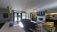 This contemporary single-floor space exudes a polished, modern aesthetic designed for comfort and socializing. The open-plan layout is anchored by a large sectional sofa in a deep charcoal hue, complemented by accent chairs in a bold mustard yellow, creating a striking contrast against the neutral gray palette throughout. A central zebra-print area rug adds a dynamic visual element underfoot, while a round wooden side table provides a refined finishing touch.

The focal wall features a sleek, tiled fireplace surround with a large flat-screen television mounted above, flanked by built-in shelving that offers both display and storage functionality. A sculptural, globe-cluster chandelier serves as an eye-catching centerpiece suspended from the smooth white ceiling, contributing to the overall upscale ambiance.

Natural light floods the interior through a generous arrangement of large windows and floor-to-ceiling glass double doors, which open to an outdoor area and provide an inviting connection to the exterior. Recessed lighting supplements the natural illumination, ensuring the space remains bright throughout the day. Dark cabinetry along the back wall adds depth and contrast, while polished countertop surfaces reflect light and enhance the sense of openness. Overall, the space balances contemporary design with a warm, welcoming atmosphere.