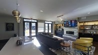 This contemporary single-floor space exudes a polished, modern aesthetic designed for comfort and socializing. The open-plan layout is anchored by a large sectional sofa in a deep charcoal hue, complemented by accent chairs in a bold mustard yellow, creating a striking contrast against the neutral gray palette throughout. A central zebra-print area rug adds a dynamic visual element underfoot, while a round wooden side table provides a refined finishing touch.

The focal wall features a sleek, tiled fireplace surround with a large flat-screen television mounted above, flanked by built-in shelving that offers both display and storage functionality. A sculptural, globe-cluster chandelier serves as an eye-catching centerpiece suspended from the smooth white ceiling, contributing to the overall upscale ambiance.

Natural light floods the interior through a generous arrangement of large windows and floor-to-ceiling glass double doors, which open to an outdoor area and provide an inviting connection to the exterior. Recessed lighting supplements the natural illumination, ensuring the space remains bright throughout the day. Dark cabinetry along the back wall adds depth and contrast, while polished countertop surfaces reflect light and enhance the sense of openness. Overall, the space balances contemporary design with a warm, welcoming atmosphere.