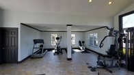 This single-floor fitness space is designed to serve as a dedicated exercise room with a practical and well-organized layout. The room features light gray walls accented by dark crown molding and base trim, creating a clean, defined aesthetic throughout. A central structural column with a matching dark base anchors the space while dividing it into functional workout zones. The flooring is a durable stone-look tile that adds texture and is well-suited for high-traffic fitness use. Multiple windows along the walls allow ample natural light to fill the room, enhancing the open, airy atmosphere. Recessed ceiling lights provide consistent overhead illumination. The room accommodates a variety of cardio equipment, including a treadmill, elliptical trainer, stationary upright bike, recumbent bike, and additional fitness machines, all arranged to allow comfortable circulation between stations. Exercise mats are positioned beneath individual machines for added stability and floor protection. The high, white ceiling contributes to the spacious feel of the room, making it a comfortable and well-appointed environment for regular fitness activity.