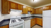 This single-story space is thoughtfully laid out to maximize functionality across its key living areas. The kitchen serves as a practical hub, featuring warm oak cabinetry that extends to the ceiling for generous storage capacity. Dark speckled countertops provide ample workspace, complemented by white appliances including a gas range, dishwasher, and refrigerator. A ventilation hood sits above the range, and a single-basin sink is positioned along the L-shaped counter layout. Light tile flooring completes the kitchen's clean, utilitarian aesthetic.

The living area offers an open, airy feel with neutral carpet flooring, smooth white walls, and a pendant light fixture overhead. A front entry door allows natural light to filter in, and the space connects directly to the kitchen, creating an efficient flow. The carpeted bedrooms are each well-proportioned, with vertical blind-covered windows that invite natural light while maintaining privacy. One bedroom features a vaulted ceiling detail that adds architectural interest and a sense of height.

The bathroom is finished with white tile flooring, a full soaking tub with an overhead shower, and a vanity topped with dark speckled countertops matching the kitchen. A small window above the tub provides natural ventilation and light, keeping the space bright and functional.