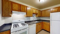 This single-story space is thoughtfully laid out to maximize functionality across its key living areas. The kitchen serves as a practical hub, featuring warm oak cabinetry that extends to the ceiling for generous storage capacity. Dark speckled countertops provide ample workspace, complemented by white appliances including a gas range, dishwasher, and refrigerator. A ventilation hood sits above the range, and a single-basin sink is positioned along the L-shaped counter layout. Light tile flooring completes the kitchen's clean, utilitarian aesthetic.

The living area offers an open, airy feel with neutral carpet flooring, smooth white walls, and a pendant light fixture overhead. A front entry door allows natural light to filter in, and the space connects directly to the kitchen, creating an efficient flow. The carpeted bedrooms are each well-proportioned, with vertical blind-covered windows that invite natural light while maintaining privacy. One bedroom features a vaulted ceiling detail that adds architectural interest and a sense of height.

The bathroom is finished with white tile flooring, a full soaking tub with an overhead shower, and a vanity topped with dark speckled countertops matching the kitchen. A small window above the tub provides natural ventilation and light, keeping the space bright and functional.