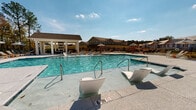 Located at 415 Curtis Pkwy SE in Calhoun, Georgia, this outdoor amenity area is designed to deliver a resort-style experience for residents. At the heart of the space is a generously sized swimming pool with crystal-clear blue water, offering varying depths clearly marked along the pool's edge. A distinctive feature of the pool is its array of in-water lounge chairs, allowing residents to relax partially submerged while soaking up the Georgia sun.

Surrounding the pool is a spacious concrete pool deck finished with a textured surface, lined with traditional lounge chairs and patio furniture positioned for optimal sun exposure and relaxation. Market umbrellas are strategically placed throughout the deck area to provide shade.

A well-appointed single-story poolside pavilion anchors the amenity area, featuring white columns, board-and-batten siding with green accent doors, and a broad covered porch equipped with ceiling fans — ideal for warm-weather comfort. The pavilion's covered outdoor area includes dining-height tables and chairs, creating a comfortable gathering space adjacent to the pool.

The facility is enclosed by a perimeter fence, providing privacy and security. The backdrop of the multi-story residential buildings and open sky enhances the expansive feel of this thoughtfully designed outdoor amenity space.