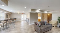 Located at 217 Binford Dr SW in Huntsville, Alabama, this single-floor community space offers a warm and functional layout designed for comfort and social interaction. The interior is finished with light hardwood-style flooring throughout, neutral taupe and beige wall tones, and recessed lighting that keeps the space bright and inviting.

The open living room is generously sized, accommodating multiple seating arrangements including a patterned sofa and upholstered accent chairs. Decorative touches such as framed artwork and potted plants add a homey, welcoming ambiance. A French door with glass pane inserts connects the living area to an adjoining room, allowing natural light to flow freely between spaces.

The dining area features multiple round tables paired with contemporary ergonomic chairs, all positioned near large windows dressed with white blinds that bring in abundant natural light and views of the surrounding greenery.

The kitchen is well-appointed with warm wood cabinetry, black appliances including a refrigerator with ice maker, a double sink, and ample counter space. A wide breakfast bar with barstool seating bridges the kitchen and the open common area, making it ideal for casual dining and social gatherings.

Overall, this space presents a clean, functional, and comfortable environment suited for community or shared use.