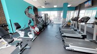This fitness center is designed to provide a comprehensive workout environment within a single, open floor layout. The space is immediately distinguished by its bold teal accent walls, exposed brick detailing, and motivational typography murals — including "You Are Stronger Than You Think" and "Sweat More" — that collectively establish an energizing and encouraging atmosphere.

The cardio section is well-stocked with Cybex treadmills, elliptical trainers, stationary bikes, and a spin bike, all arranged to maximize floor space and allow for easy movement between machines. Large windows along one wall flood the area with natural light and provide views of the surrounding exterior, enhancing the open feel of the space. Full-length mirrors along one side further amplify the sense of spaciousness and allow users to monitor their form.

A dedicated strength training area features a multi-use cable machine, a weight bench with a barbell rack, and a dumbbell rack stocked with a range of free weights. An Everlast speed bag mounted to the exposed brick wall adds a boxing element to the facility. Ceiling fans and recessed panel lighting ensure consistent comfort and visibility throughout. Durable rubber flooring covers the entire space, providing safety and support across all workout zones. A wall-mounted television is also present for entertainment during cardio sessions.