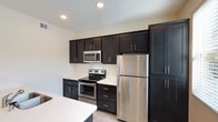 Located at 10785 Pomerado in San Diego, Unit #402 is a well-appointed single-floor residence designed with a clean, contemporary aesthetic throughout. The kitchen is a standout feature, showcasing dark espresso shaker-style cabinetry extending to the ceiling for ample storage, crisp white countertops, and a full suite of stainless steel appliances including a refrigerator, range, and microwave. A peninsula with bar seating and a double-basin sink adds both functionality and a casual dining option, while light wood-style flooring ties the space together. The kitchen opens seamlessly into a generously sized living area carpeted in neutral tones and bathed in natural light from large windows fitted with vertical blinds and shutters. A half bathroom off the main living area provides added convenience for guests, featuring a pedestal sink and mirror. The bedroom is a comfortable retreat with plush carpet, a dedicated walk-in closet, and access to an additional closet for extra storage. The full bathroom includes dark cabinetry with a white countertop vanity, a separate water closet, and a walk-in shower. Recessed lighting throughout the unit contributes to a bright, modern ambiance, while the neutral color palette creates a versatile and inviting atmosphere.