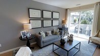 This single-floor space showcases a refined and contemporary aesthetic, centered around a well-appointed living area that blends comfort with sophisticated design. The living room is anchored by a sleek gray sofa adorned with a mix of patterned and neutral throw pillows, complemented by a warm-toned area rug featuring a subtle botanical motif. A striking gallery wall displays a symmetrical arrangement of six black-framed artworks, adding a curated, gallery-like quality to the space. Natural light flows generously through a large sliding glass door that opens directly onto an outdoor patio, offering views of an exterior seating area surrounded by lush greenery. The room is further enhanced by a dark wood coffee table with a metal frame, a coordinating side table with a classic pedestal base, and warm ambient lighting provided by table lamps with crisp white shades. Decorative accents, including an orchid arrangement and sculptural pieces, add organic texture and personality. The neutral gray wall tones create a calm, versatile backdrop that ties the entire space together, making it both inviting and visually cohesive.