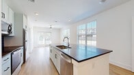 This single-floor residence offers a clean, contemporary design throughout, characterized by crisp white walls, light wood-look flooring, and an abundance of natural light streaming through multiple windows fitted with horizontal blinds. The heart of the home is an open-concept kitchen anchored by a generously sized island topped with dark granite countertops and equipped with an undermount sink and pull-down faucet. White shaker-style cabinetry lines the walls, complemented by a slate tile backsplash and a full suite of stainless steel appliances, including a refrigerator, range, microwave, and dishwasher. The kitchen flows seamlessly into the living area, which features a ceiling fan and access to the outdoors via French doors, further enhancing the bright and airy atmosphere. The bedrooms are carpeted for comfort, each with a ceiling fan and windows that invite natural light. One room opens directly to an adjacent space, offering flexible use. The bathroom features a single-sink vanity with dark granite countertops, grey cabinetry, a large mirror with overhead lighting, and a glass-enclosed walk-in shower set on wood-look flooring, creating a clean and functional design. Overall, this residence combines modern finishes with a practical, open layout.