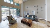 This single-floor amenity space is thoughtfully designed to offer a variety of social and functional areas within a cohesive, contemporary aesthetic. The grand lobby serves as an impressive entry point, featuring soaring double-height ceilings, large floor-to-ceiling glass doors that invite abundant natural light, and striking built-in vertical botanical art panels flanking the main corridor entrance. Warm hardwood-style flooring flows consistently throughout, unifying the space.

The kitchen and bar area is a central highlight, outfitted with expansive granite countertops, dark espresso cabinetry, a stainless steel dishwasher, an undermount sink, and a row of bold matte black pendant lights overhead. Bar-height seating lines the granite island, creating an inviting gathering spot. Multiple wall-mounted televisions enhance the entertainment-friendly atmosphere.

Adjacent lounge areas feature distinct personalities: one showcases a textured mosaic accent wall paired with a sectional sofa, while another offers a built-in white bookcase, a stone-clad fireplace wall, and corridor access to additional rooms. A dining nook with a round table and glass French doors opens directly to an outdoor balcony with open views. Throughout, warm neutral tones, curated wall art, and recessed lighting contribute to a polished, welcoming ambiance.