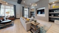This thoughtfully designed single-floor amenity space blends contemporary style with functional versatility across several distinct areas. Light-toned large-format tile flooring flows throughout, creating a cohesive and airy atmosphere.

The welcoming entryway features soaring ceilings, abstract aqua-toned wall art, and ample natural light from floor-to-ceiling windows with draping curtains. A cozy seating nook nearby showcases a teal accent wall adorned with circular mirrors, complemented by plush navy wingback chairs and open wooden shelving.

The central lounge area is anchored by a generous sectional sofa, a circular ring chandelier overhead, and a built-in wood-paneled media wall with integrated shelving. Bold patterned drapes frame the glass doors that open to an outdoor terrace, filling the space with natural light.

The kitchen bar features a long waterfall-edge countertop with sleek wood cabinetry, striking navy vertical tile backsplash, open floating shelves, and dramatic black-and-gold pendant lighting — ideal for casual gatherings.

The billiards room offers a moody, sophisticated atmosphere with deep navy walls, a large pool table centered on an abstract area rug, and a wall-mounted television. The business center provides a bright, teal-walled workspace equipped with computers and ample desk seating beneath globe pendant lights. Double glass doors lead to an additional fitness or activity room beyond.