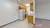 This single-floor space offers a straightforward, functional layout organized around a central corridor that connects the main living areas. The living area is generously sized, featuring neutral beige carpeting, crisp white walls, and wood baseboards that run throughout. Natural light filters in through multiple windows, including a pair of windows that bring in views of surrounding greenery, contributing to a calm and open atmosphere. A baseboard heating system lines the perimeter, providing efficient climate control.

The kitchen is compact yet well-equipped, featuring warm honey-toned wood cabinetry that extends to the upper walls for ample storage. White appliances, including a refrigerator and electric range, are neatly arranged along the walls, while a peninsula with a built-in sink adds a practical workspace. The kitchen floor is finished with light-toned tile, creating a clean, easy-to-maintain surface.

The bedroom is a comfortable, carpeted room with white walls and a full-width sliding closet fitted with wood-panel doors, offering substantial storage space.

The bathroom is neatly appointed with white tile walls, a tub-shower combination, a white vanity with cabinet storage, a mirrored medicine cabinet, and light travertine-style tile flooring, giving the room a clean and orderly appearance.