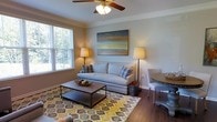 This single-floor space offers a thoughtfully designed interior that balances comfort and style. The living area serves as the heart of the home, featuring rich hardwood flooring that adds warmth and character throughout. A ceiling fan with a light fixture anchors the room from above, providing both practical airflow and ambient lighting. Crown molding along the upper walls adds a refined architectural detail that elevates the overall aesthetic.

Natural light flows generously through a large window fitted with blinds, offering views of lush greenery outdoors. The living space is furnished with a classic nailhead-trim sofa accented with decorative throw pillows, complemented by a sturdy industrial-style wooden coffee table. A vibrant geometric-patterned area rug in warm yellows, greens, and grays grounds the seating area and adds a pop of color against the neutral walls.

A large landscape-style artwork depicting a serene horizon hangs prominently on the main wall, reinforcing the calm, coastal-inspired ambiance of the room. Floor lamps on either side of the sofa provide soft, layered lighting. Adjacent to the living area, a glimpse of a compact dining nook with a round wooden table and upholstered seating rounds out this inviting, cohesive living space.