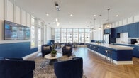 Located at 12525 Oldehurst Place in Charlotte, NC, this single-floor community clubhouse showcases a striking contemporary interior defined by a bold navy and white color palette. The herringbone-patterned hardwood flooring runs throughout, lending a sense of cohesion and sophistication to the entire space.

The kitchen is a standout feature, outfitted with deep navy shaker-style cabinetry, white subway tile backsplash, stainless steel appliances, and an expansive white quartz island accommodating multiple bar stools. Elegant globe pendant lights hang overhead, adding warmth and visual interest to the space.

Adjacent to the kitchen, a lounge area provides a comfortable gathering space furnished with plush navy velvet armchairs arranged around a round coffee table atop a patterned area rug. A wall-mounted television, decorative geometric wall paneling, and cluster pendant lighting complete this inviting setting.

A separate dining area is bathed in natural light from floor-to-ceiling windows that frame views of an outdoor pool and the surrounding community. Multiple café-style tables and chairs are arranged throughout, making it an ideal spot for casual gatherings or remote work.

Throughout the space, white vertical wall paneling with a navy wainscot accent creates a refined, layered aesthetic, while recessed lighting ensures the entire clubhouse remains bright and welcoming at all times.