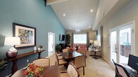 The clubhouse at 5427 Lake Murray in La Mesa is a thoughtfully designed single-story community space that blends comfort with style. The interior is immediately distinguished by its dramatic vaulted ceilings with exposed beams, which lend an airy, spacious quality to the open-plan layout. A bold teal accent wall adds a vibrant pop of color, complementing the neutral beige tones found throughout the space.

The layout flows seamlessly from a defined dining area, furnished with a rich dark wood table and patterned upholstered chairs, into a relaxed lounge area. The lounge features coral-toned accent chairs and a neutral sofa arrangement, creating an inviting setting for casual conversation or community events. Recessed lighting and a decorative globe chandelier provide warm, layered illumination throughout.

Natural light enters through large windows dressed with white blinds and gray curtain panels, brightening the interior while maintaining privacy. A wall-mounted flat-screen television serves the lounge area, adding a practical amenity for residents. Potted greenery and carefully chosen décor accessories contribute to the welcoming, well-appointed ambiance of this community gathering space.