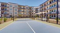 Located at 17120 North 53rd Avenue in Glendale, Arizona, this outdoor amenity space centers around a well-maintained pickleball court set within a modern multi-story residential community. The court features a regulation-size playing surface with a blue and green color scheme, clearly marked boundary lines, and a standard net, all enclosed by a black chain-link fence with a dedicated entry gate. Flanking the court on multiple sides are the community's contemporary multi-story buildings, characterized by neutral stucco exteriors, large windows, and private balconies that overlook the activity below. Adjacent to the court, a covered pergola structure provides shaded seating, making it an ideal spot for spectators or social gatherings. String lights are strung between posts, adding a welcoming ambiance for evening use. The surrounding landscape incorporates desert-adapted plantings, mulched beds, and young trees that frame the space with a clean, manicured aesthetic. A stretch of open lawn is also visible nearby, complementing the recreational atmosphere. Tall light poles ensure the court is well-lit for use beyond daylight hours. The open Arizona sky above bathes the entire area in natural sunlight, reinforcing the active, outdoor lifestyle that defines this community amenity.