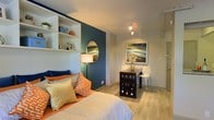 This single-floor studio is thoughtfully designed to maximize both function and style within a compact footprint. The space immediately draws attention with a striking teal accent wall that serves as a dramatic backdrop to the sleeping area, creating a bold focal point that anchors the room. Built-in white open shelving lines one wall, offering practical storage and display space for decorative items, books, and personal touches, adding depth and character to the layout.

Light wood-toned flooring runs throughout, lending warmth and visual continuity to the open-plan interior. The sleeping area features a generously sized bed dressed in layered neutral and orange accent pillows, complemented by a classic floor lamp that adds soft, ambient lighting. A round mirror mounted on the teal wall enhances the sense of space and light.

A compact dining or workspace nook is positioned near a framed landscape artwork, accompanied by a dark wine storage cabinet and a sleek bar-height chair — a clever use of space that combines utility with aesthetic appeal. Recessed track lighting on the ceiling provides focused illumination throughout. The overall ambiance is modern, cozy, and well-curated, making efficient use of every corner while maintaining a welcoming and stylish atmosphere.