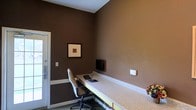 This single-floor space is designed as a focused, functional workspace with a clean and professional atmosphere. The room is finished with rich, dark brown walls that create a warm and focused environment, complemented by crisp white trim along the ceiling and baseboards for a polished contrast.

A standout feature of the space is the long, built-in countertop workstation that spans a generous stretch of the wall, providing ample surface area for productivity. The workstation is equipped with a keyboard, a wall-mounted flat-screen monitor, and an ergonomic office chair, making it well-suited for computer work or administrative tasks.

A framed piece of artwork adorns one of the dark walls, adding a subtle decorative touch that softens the otherwise utilitarian design. The room also includes a wall-mounted control panel or outlet, suggesting thoughtful integration of technology and utilities throughout.

Access to the space is provided through a secure door with a visible latch mechanism, hinting at a professional or shared-use environment. The overall ambiance is intimate yet purposeful, offering a quiet and distraction-free setting ideal for focused work or a business center-style use.
