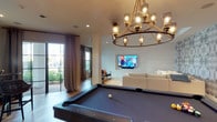This single-floor space is thoughtfully designed as a sophisticated entertainment retreat, blending coastal-modern aesthetics with functional leisure areas. The layout flows seamlessly between distinct zones, each with its own character while maintaining a cohesive design language.

The entertainment lounge serves as a central focal point, anchored by a large wall-mounted flat-screen television recessed into white shiplap paneling. A generously sized sectional sofa in neutral tones faces the screen, complemented by a striking live-edge wood coffee table and a soft area rug. A bold, abstract patterned wallcovering in muted grey and blue tones adds dramatic texture to the adjacent wall.

The games area features a full-sized pool table beneath an elegant multi-arm chandelier with glass cylindrical shades, adding warmth and visual interest overhead. Natural light filters through black-framed glass doors and windows dressed with floor-length linen drapes, connecting the interior to lush greenery outside.

A dedicated bar area offers a sleek stone countertop with woven rattan barstools, custom cabinetry, and the same eye-catching wallcovering found in the lounge. A bistro-style high table with a round reclaimed-wood top and oversized circular mirror creates an intimate secondary seating nook. Light hardwood flooring runs throughout, tying the entire space together.