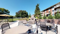 This single-floor community space offers a thoughtfully designed combination of outdoor amenities and functional interior facilities. The expansive outdoor patio is finished with a smooth concrete surface and outfitted with multiple seating arrangements, including round tables and metal chairs suited for group gatherings. A built-in masonry fireplace serves as a central focal point of the patio, framed by raised planter beds and surrounded by lush, mature trees that provide natural shade and greenery. The open-air setting enjoys abundant natural light under clear skies, with views of the broader landscaped grounds and a multi-story residential building visible in the background. A fenced recreational area, likely a tennis or sport court, can be seen in the distance, adding to the range of outdoor amenities available.

The interior restroom facility is clean and efficiently organized, featuring multiple stainless steel sink basins with chrome faucets and glass cup holders mounted above warm wood-toned cabinetry below. The under-sink areas are fitted with open shelving providing practical storage. The walls are finished in a neutral tone, contributing to a tidy and functional atmosphere. Together, the outdoor and indoor elements of this space create a comfortable, community-oriented environment designed for both relaxation and convenience.