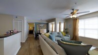 This single-floor space offers a thoughtfully designed open-concept layout that seamlessly connects the living, dining, and sleeping areas. The living room serves as the heart of the home, featuring warm hardwood-style flooring, a ceiling fan with integrated lighting, and generous natural light streaming through windows dressed in rich golden drapes. The seating area is furnished with a neutral sectional sofa accented by yellow and teal throw pillows, complemented by a pair of light blue armchairs, creating a cohesive and inviting color palette throughout. Adjacent to the living room, the dining area is distinguished by a bold teal accent wall adorned with a decorative floral art piece, anchored by a dark dining table beneath a classic pendant light fixture. The bedroom, visible through a connecting hallway, continues the warm tones with hardwood-style flooring and soft ambient lighting. Throughout the space, the interplay of neutral walls, vibrant accent colors, and carefully selected furnishings creates a balanced and welcoming ambiance. The open flow between rooms enhances the sense of spaciousness, making this a comfortable and visually appealing single-level residence.