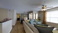 This single-floor space offers a thoughtfully designed open-concept layout that seamlessly connects the living, dining, and sleeping areas. The living room serves as the heart of the home, featuring warm hardwood-style flooring, a ceiling fan with integrated lighting, and generous natural light streaming through windows dressed in rich golden drapes. The seating area is furnished with a neutral sectional sofa accented by yellow and teal throw pillows, complemented by a pair of light blue armchairs, creating a cohesive and inviting color palette throughout. Adjacent to the living room, the dining area is distinguished by a bold teal accent wall adorned with a decorative floral art piece, anchored by a dark dining table beneath a classic pendant light fixture. The bedroom, visible through a connecting hallway, continues the warm tones with hardwood-style flooring and soft ambient lighting. Throughout the space, the interplay of neutral walls, vibrant accent colors, and carefully selected furnishings creates a balanced and welcoming ambiance. The open flow between rooms enhances the sense of spaciousness, making this a comfortable and visually appealing single-level residence.